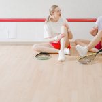 Two women sitting on a wooden floor with tennis rackets, smiling and chatting.